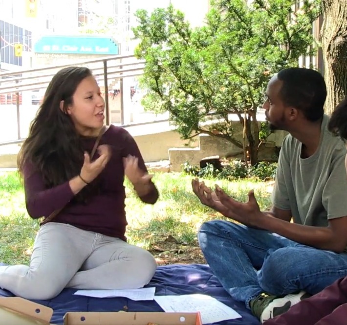 a young man and a young woman sitting on the ground outside signing in conversation