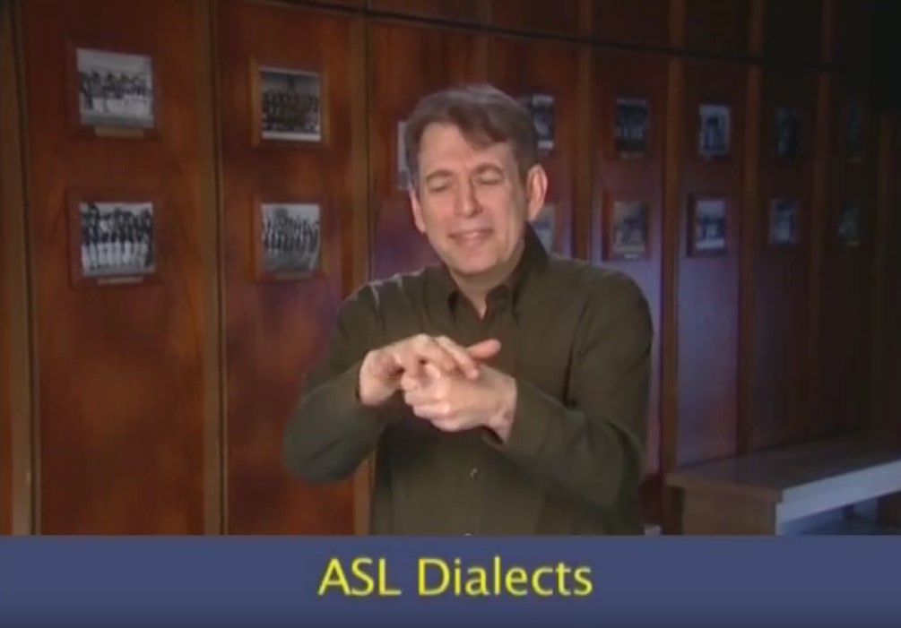 man signing in a hallway with many old photos. On the bottom of the image it says "ASL Dialects"